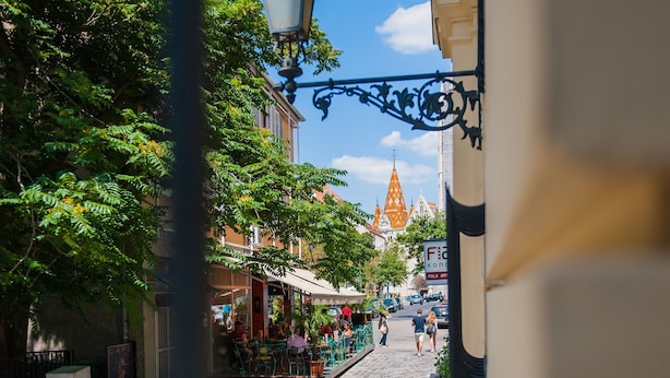 Una calle adoquinada bordeada de cafés y tiendas, con una imponente aguja de iglesia al fondo y un cielo azul despejado.