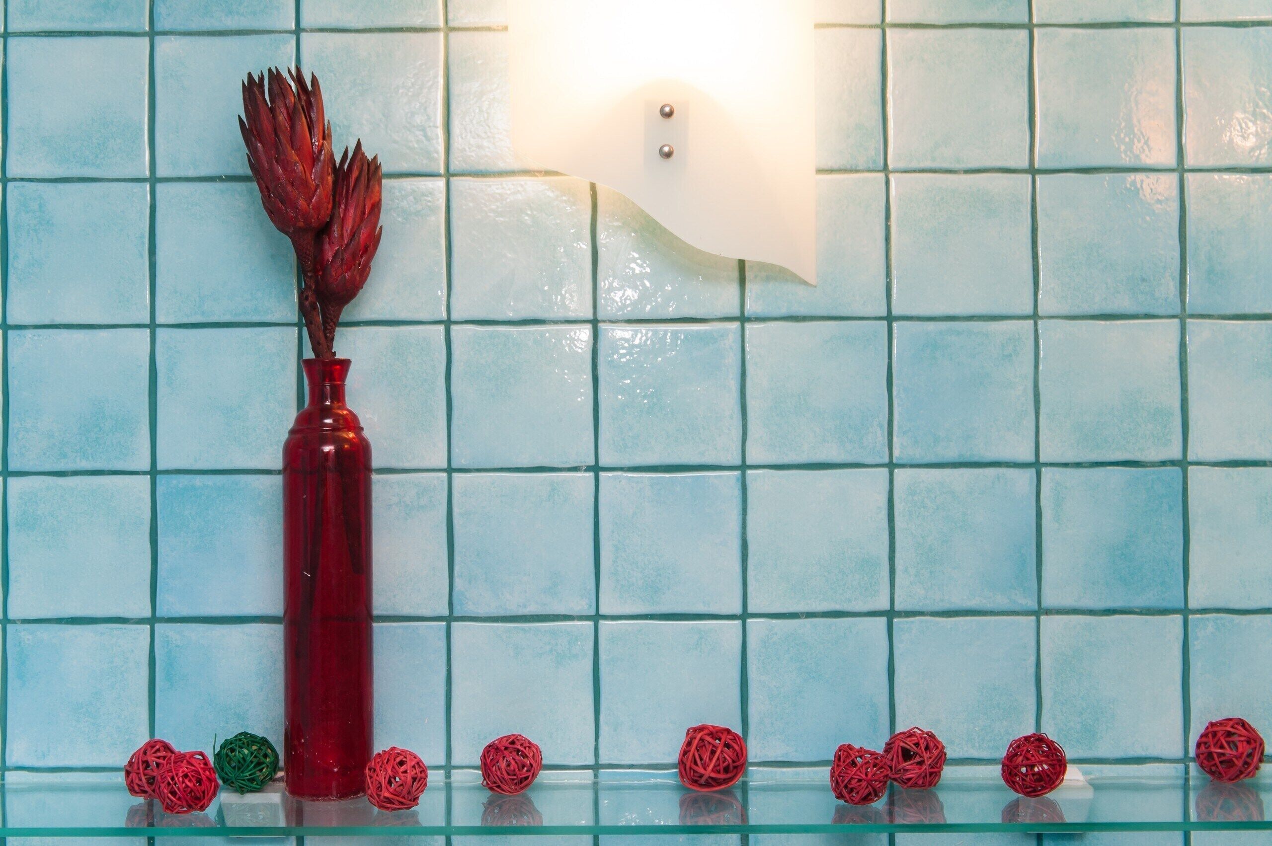 Ein Badezimmer mit einer gefliesten Wand, einer roten Vase mit getrockneten Blumen und mehreren roten und grünen Dekokugeln auf einem Glastisch.
