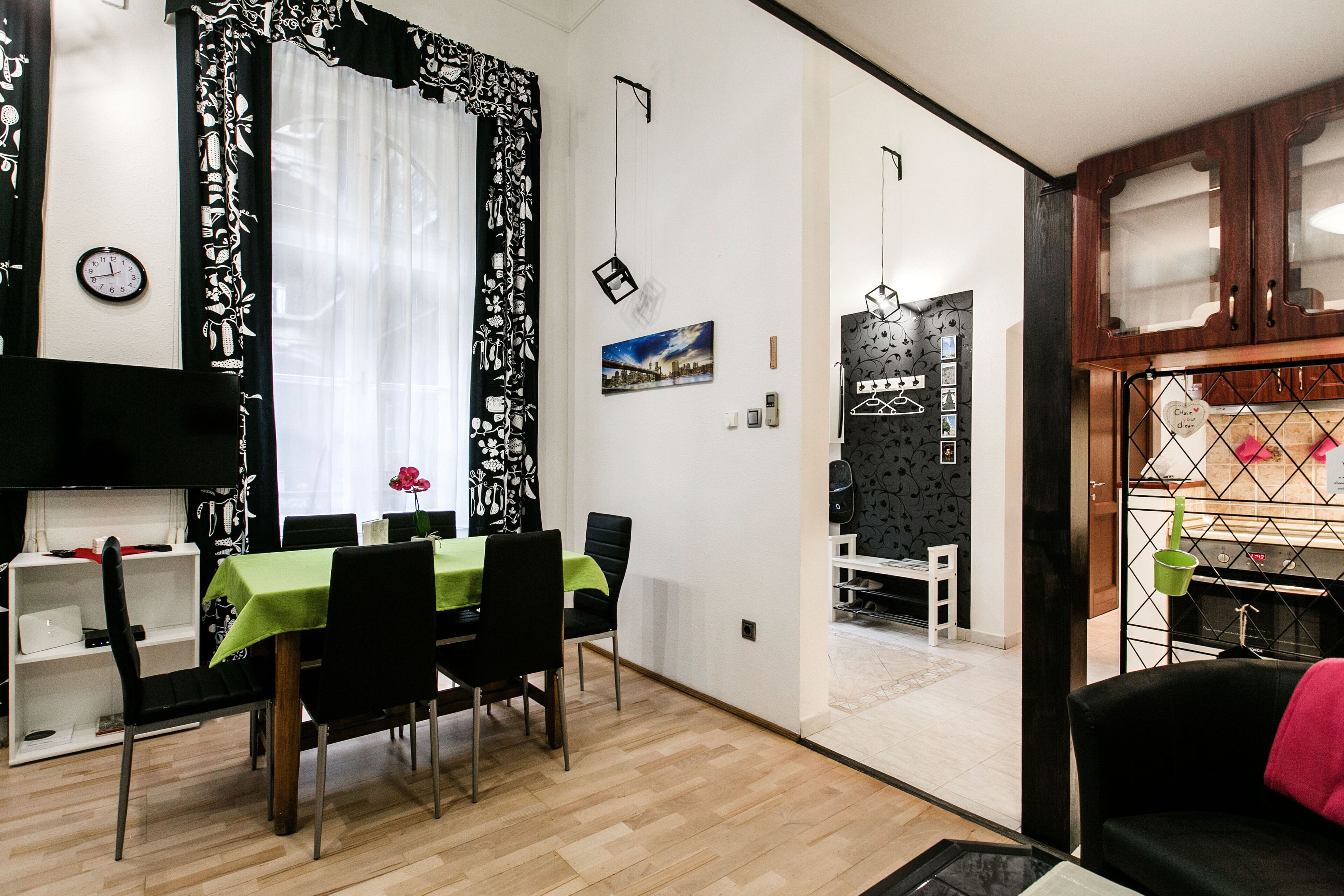 Un coin repas avec une table dressée pour quatre personnes, une horloge au mur et une cuisine visible à travers une porte ouverte.