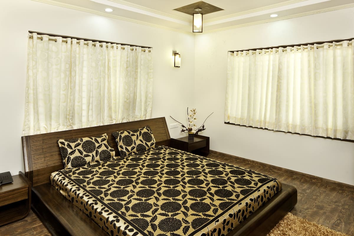 Un dormitorio con una cama grande, cabecero de madera oscura y ropa de cama estampada. Hay dos mesitas de noche con lámparas y un jarrón de flores. Las paredes son blancas y tiene una lámpara en el techo.
