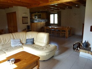 Uma sala de estar espaçosa com um amplo sofá modular bege, uma mesa de centro de madeira, e um cão pequeno. Cozinha com armários de madeira e uma área de refeições com mesa e cadeiras são visíveis ao fundo.