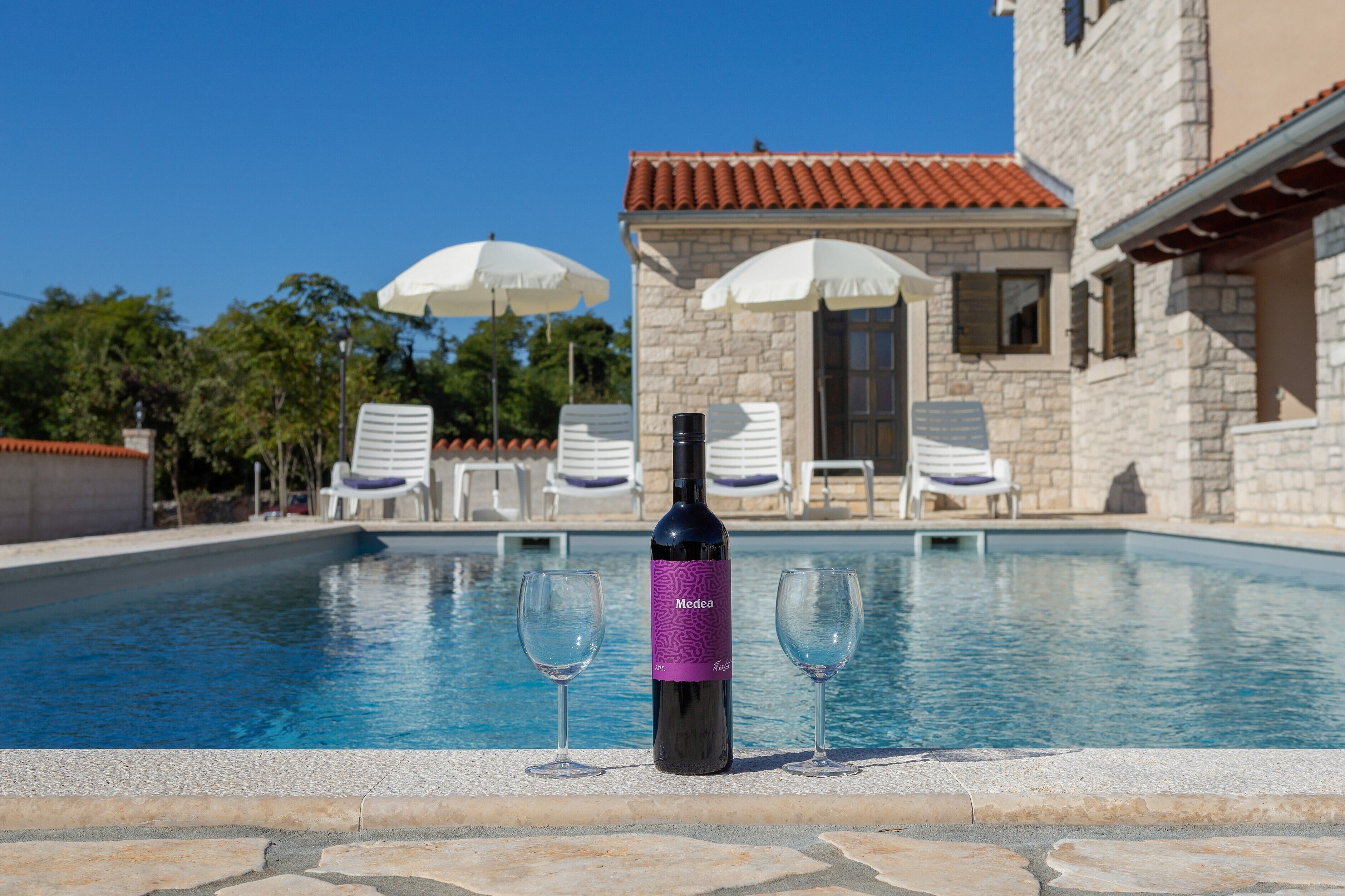 A pool area with a bottle of wine and two glasses, a stone building with a tiled roof, and white umbrellas.