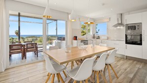 A modern kitchen with white cabinets, a wooden dining table, and a view of the sea.