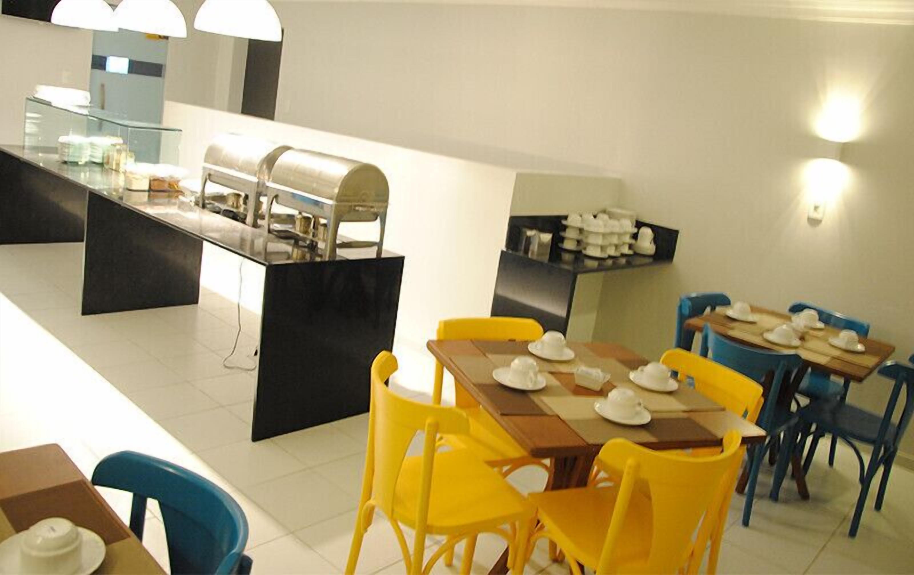 Uma área de refeições com cadeiras amarelas, uma mesa de madeira e um balcão de buffet com alimentos.