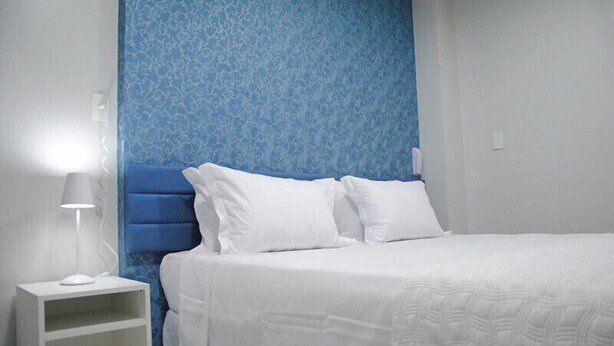 Un lit bien fait, avec une literie blanche, une tête de lit bleue et une table de chevet sur laquelle se trouve une lampe.