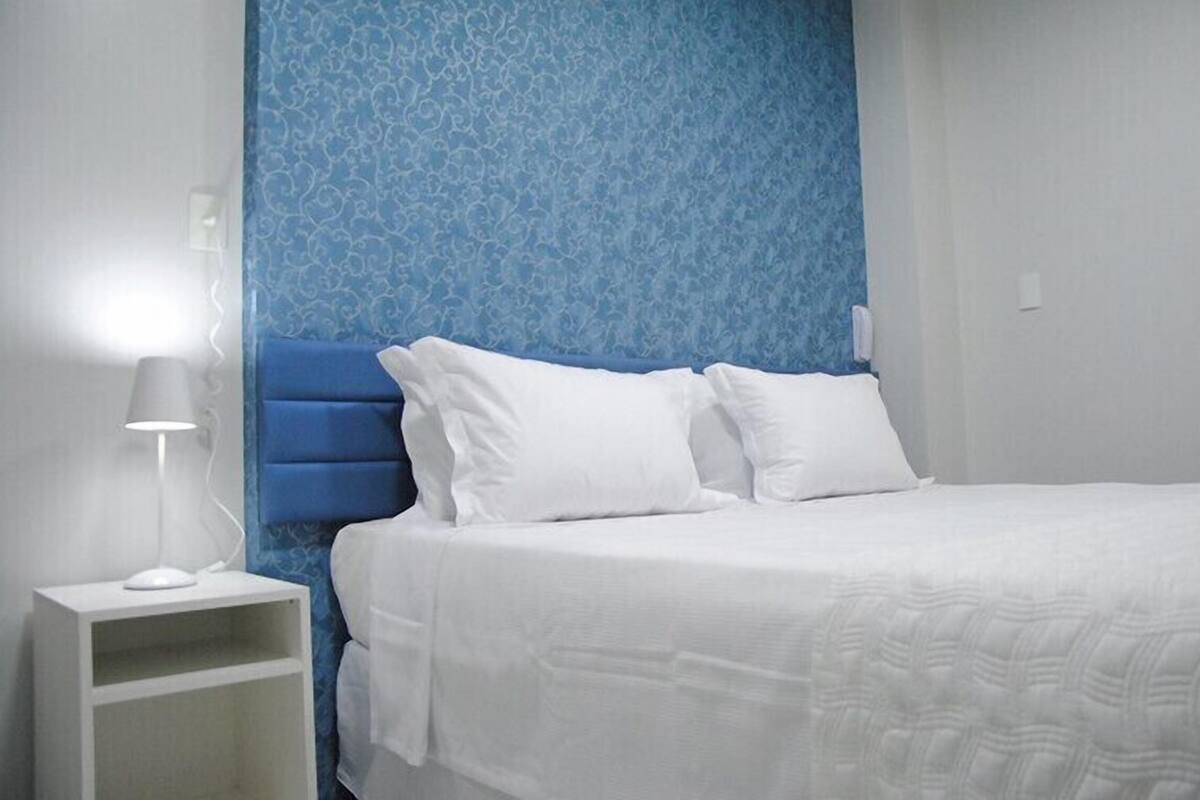 A neatly made bed with white bedding, a blue headboard, and a bedside table with a lamp.