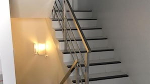 Un escalier doté d’une rampe métallique et de marches blanches, menant à un palier où est fixée une applique murale.