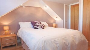 Un dormitorio con techo inclinado, una cama con ropa de cama blanca, dos lámparas de noche y una mesita de noche de madera con cajones.