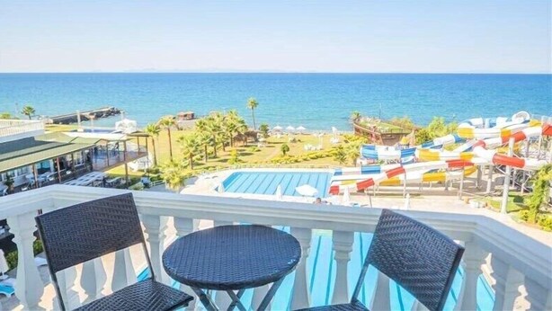 A balcony with wicker chairs and a small table, overlooking a pool area with umbrellas and lounge chairs, and a view of the sea.