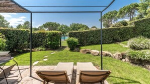 Une terrasse avec deux fauteuils de jardin en bois et une table, entourée d’un jardin bien entretenu, agrémenté de haies et d’un chemin en pierre.