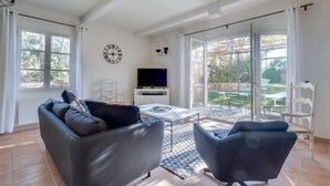Un salon spacieux comprenant un canapé, des fauteuils, un téléviseur et une grande fenêtre donnant sur l’extérieur.