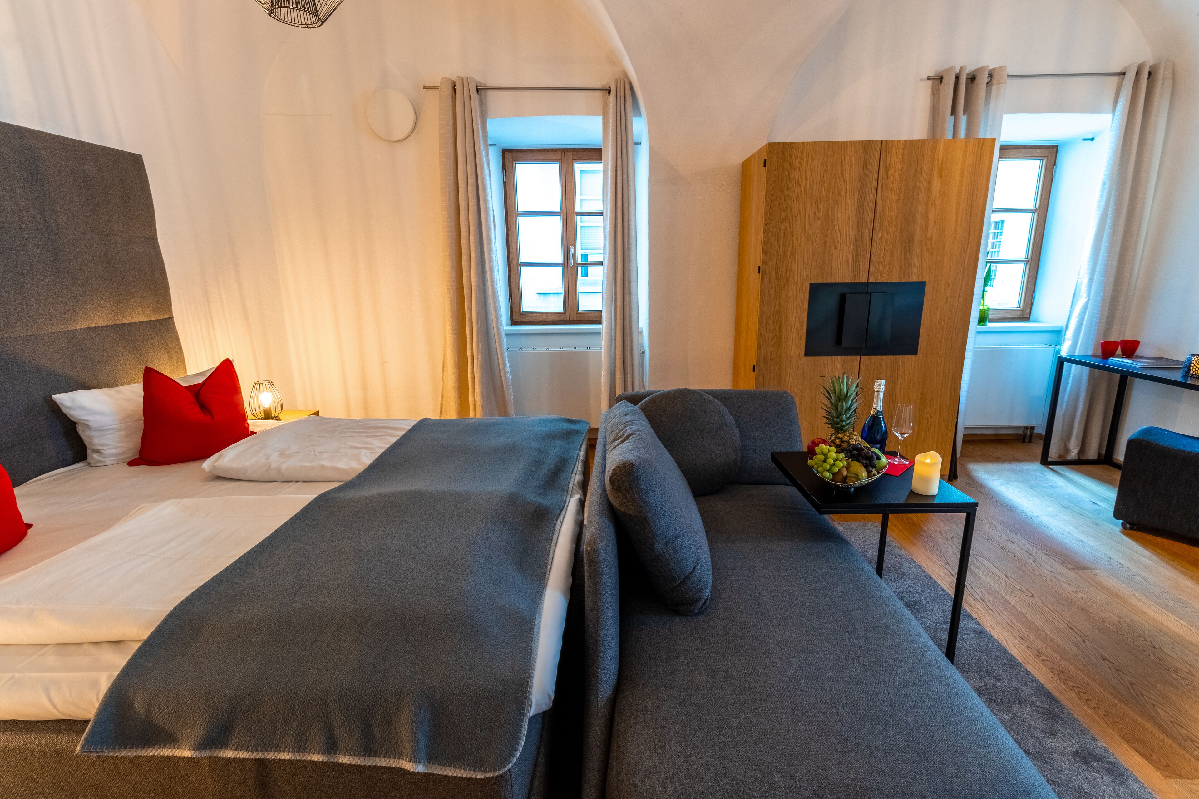 Quarto de hotel moderno com uma cama grande, um sofá, uma televisão e uma pequena mesa com uma cesta de frutas e velas.