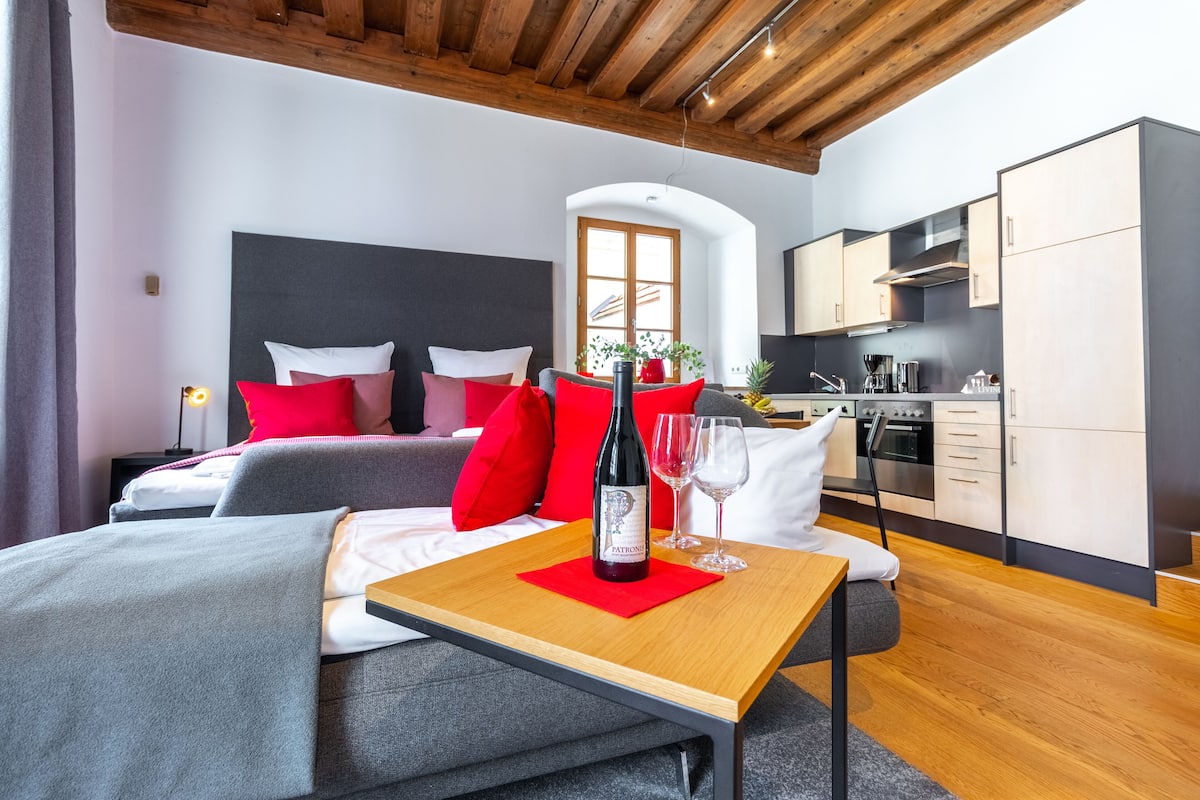 A modern room with a bed, a table with a bottle and glasses, a kitchen area, and a window with plants.