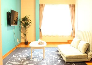 A living room with a sofa, a small table, a potted plant, and a TV mounted on the wall.
