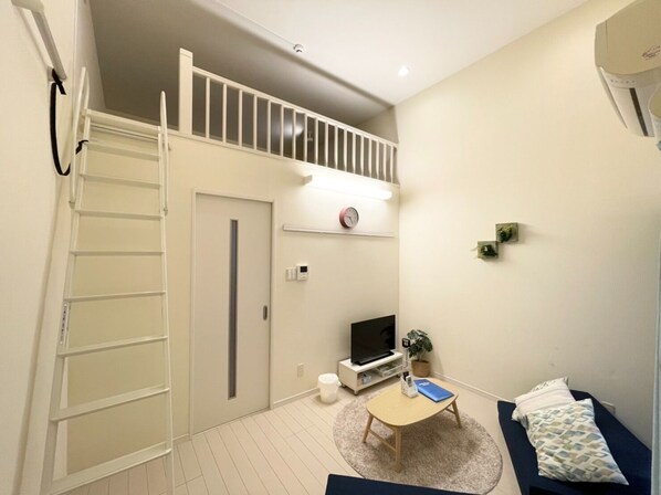 A compact living space with a loft bedroom, a small seating area with a table, a TV, and a potted plant.