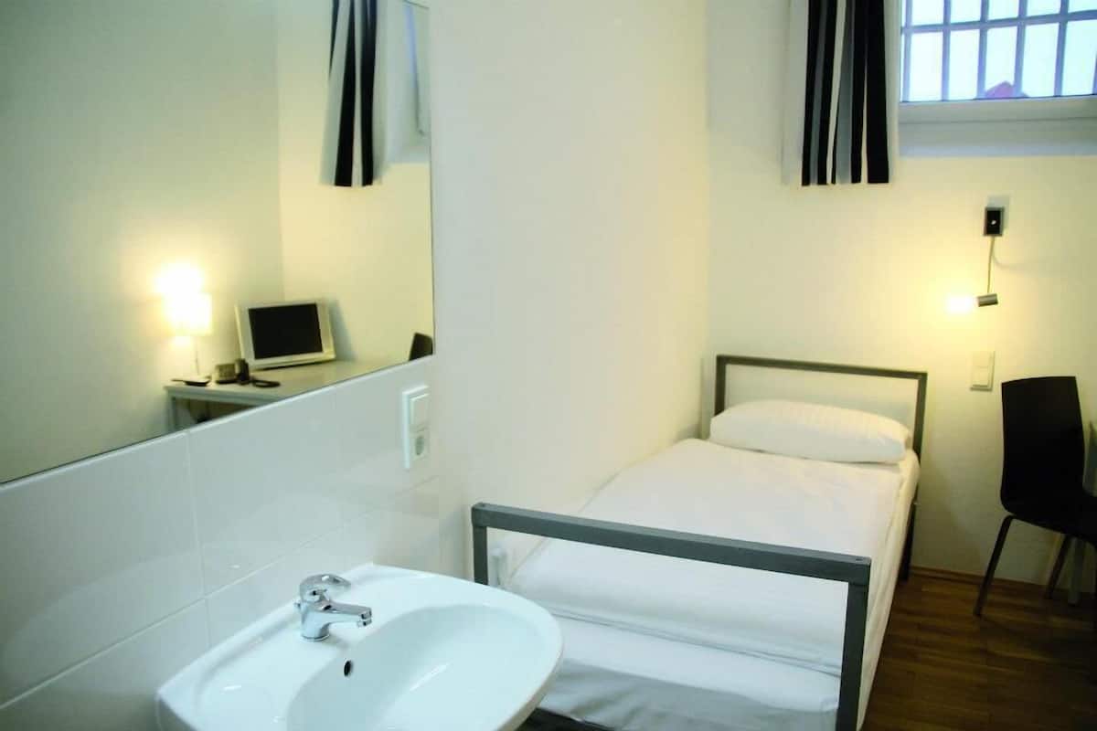 Ein kleines, sauberes Zimmer mit einem Einzelbett, einem Nachttisch mit Lampe, einem Sessel und einem großen Spiegel.