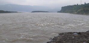 A river with turbulent water, a rocky shore, and distant mountains. - Hotel Aashirwad (Reasi)