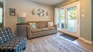 A cozy living room with a sofa, a small table with a lamp, and a patterned ottoman. - Family Tradition - Duck Landing, Oceanside, 130 Yds to Beach, Pool, Hot Tub, Tiki Bar (Duck)