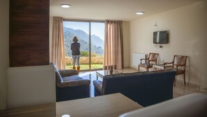 Una habitación con un amplio ventanal corredero que da a un balcón con vistas a la montaña, un televisor de pantalla plana montado en la pared y una zona de estar con mesa y sillas.