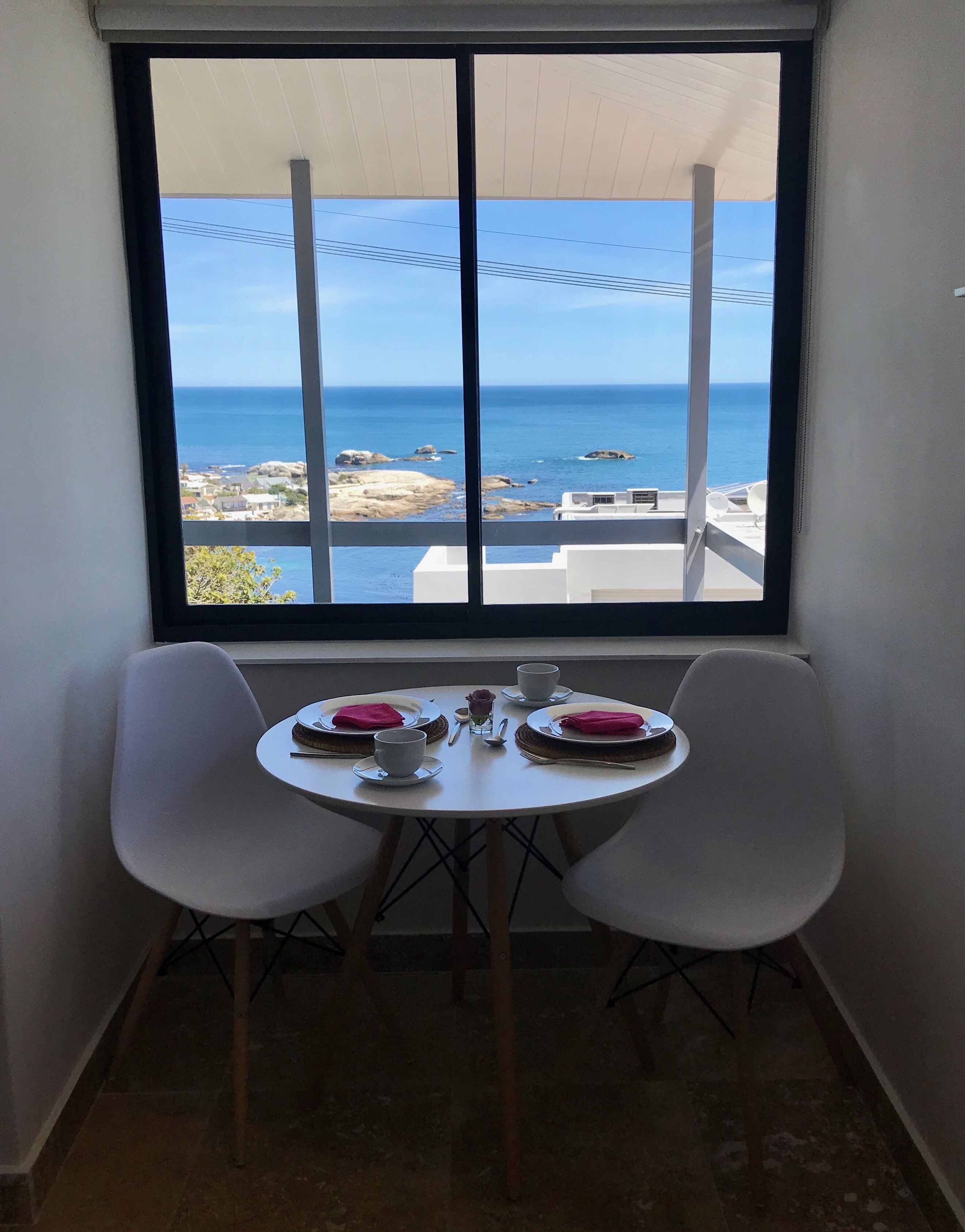 A small room with a window overlooking the sea. There is a table with two chairs and a vase of flowers.