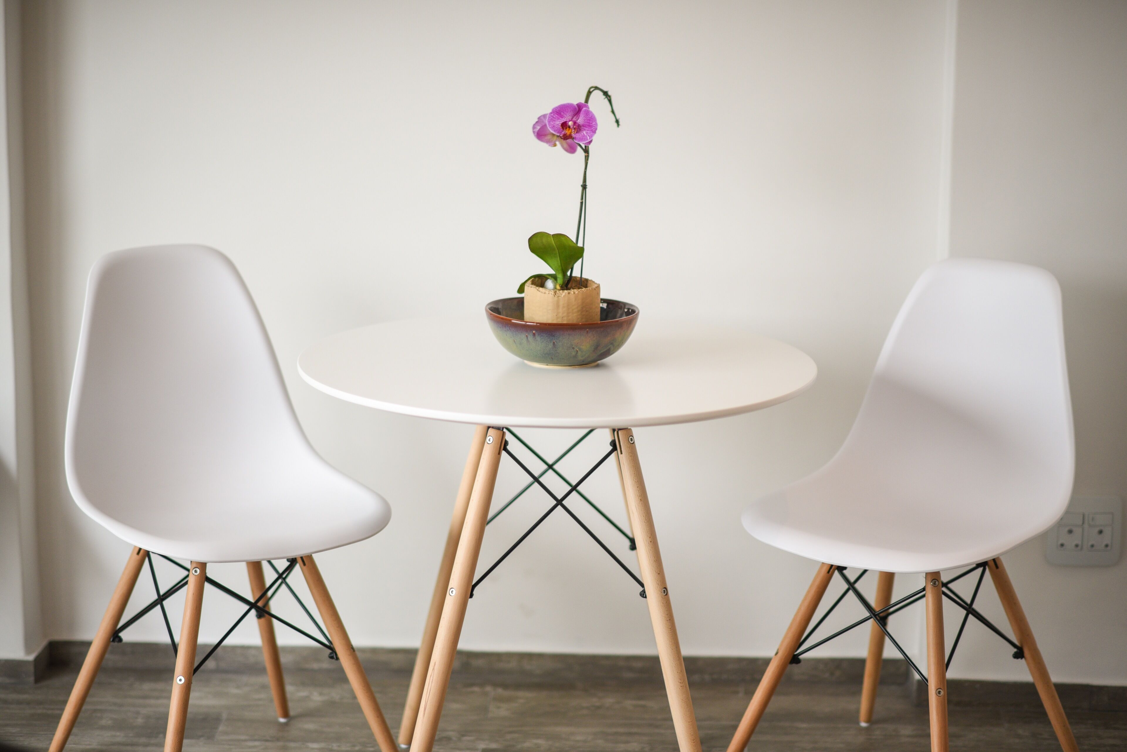 A minimalist dining setup with two white chairs and a small table, featuring a single orchid in a ceramic bowl.