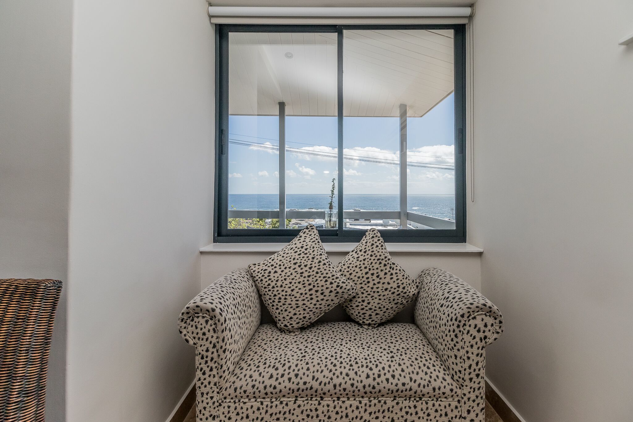 Une pièce d’angle avec un canapé et deux coussins à motifs, une grande fenêtre donnant sur la mer et un bâtiment situé au loin.