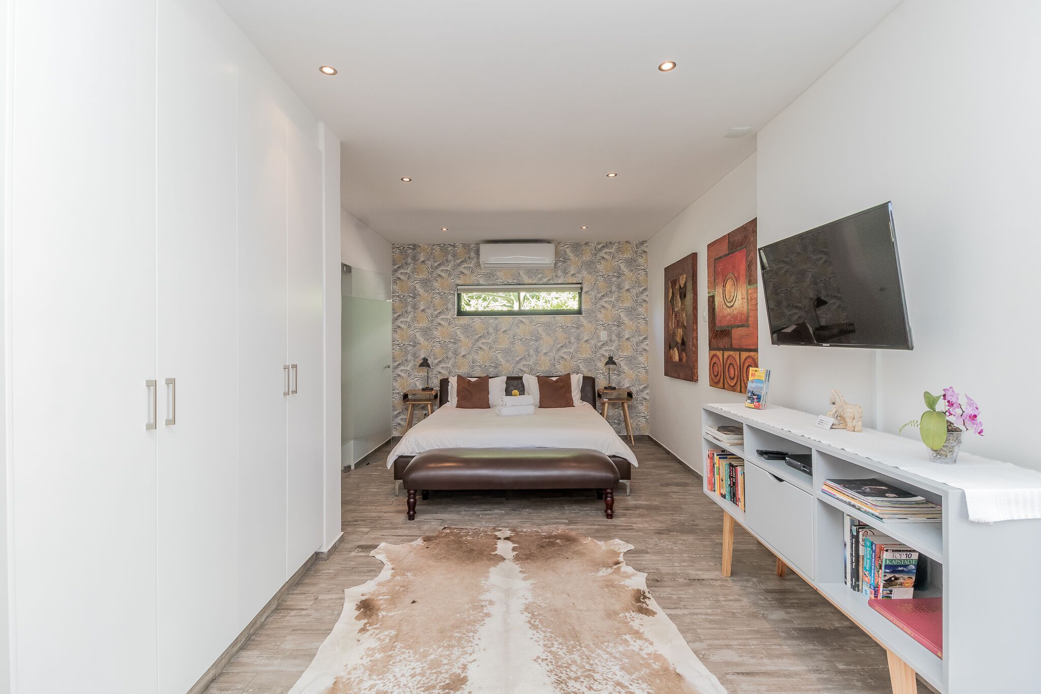 A bedroom with a large bed, a TV mounted on the wall, a bookshelf, and a window with a view.