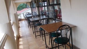 A corridor with multiple tables and chairs, leading to an outdoor area with a red roof and greenery. - Mash Park Hotel Kisii (Kadongo)