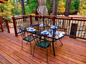 A wooden deck with a glass table set for dining, surrounded by metal chairs. - Tuckett Inn | Walk to Town + Fenced Yard (Idyllwild)