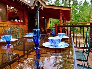 A table set with blue glassware and white plates with blue patterns, a metal chair, and a wooden deck with a railing. - Tuckett Inn | Walk to Town + Fenced Yard (Idyllwild)