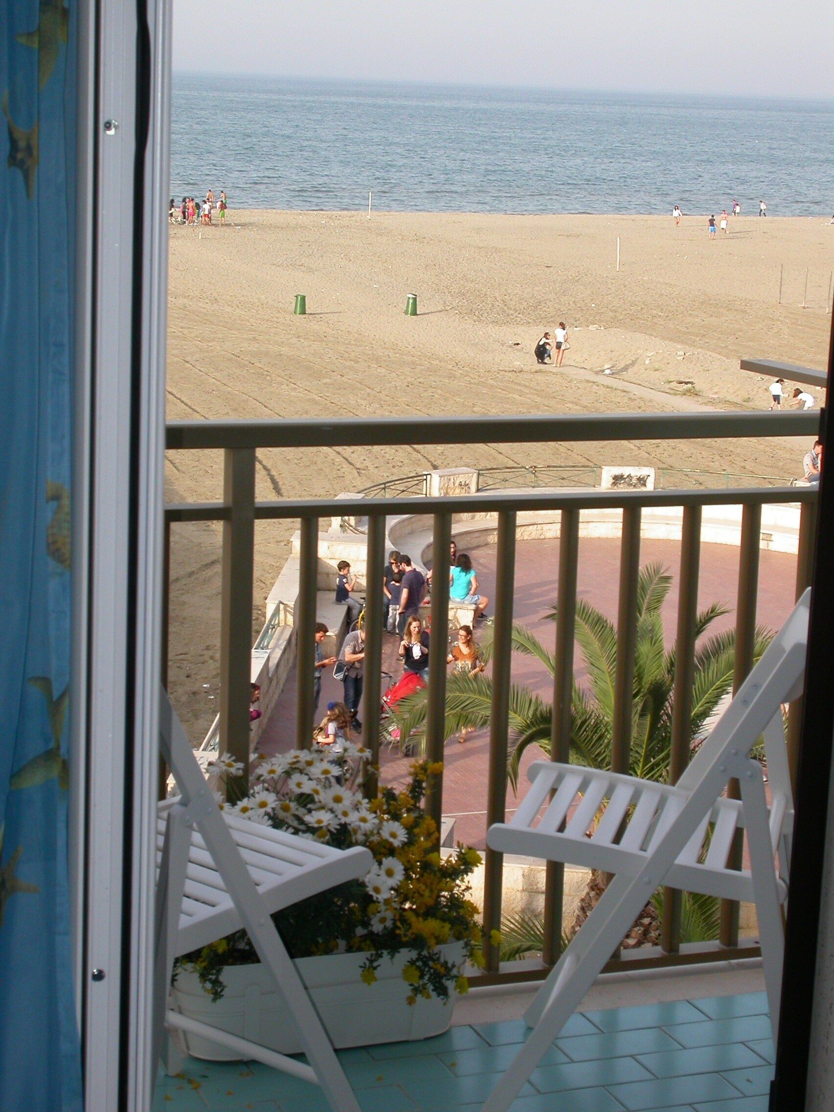 A balcony with white chairs and a flower pot, overlooking a beach with people and a sandy area.