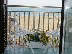 Un balcón con dos sillas blancas y un arreglo floral, con vista a una playa arenosa donde se ven personas a lo lejos.
