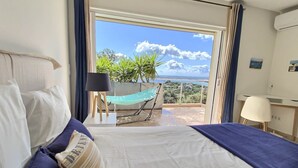 Quarto de hotel com cama, escrivaninha, cadeira, rede e varanda com vista para o verde e o céu.