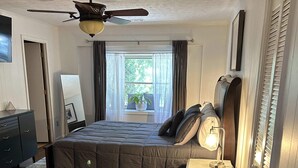 Une chambre à coucher comprenant un lit, une table de chevet, une chaise, un ventilateur de plafond, une fenêtre avec des rideaux et une vue sur la verdure à l’extérieur.