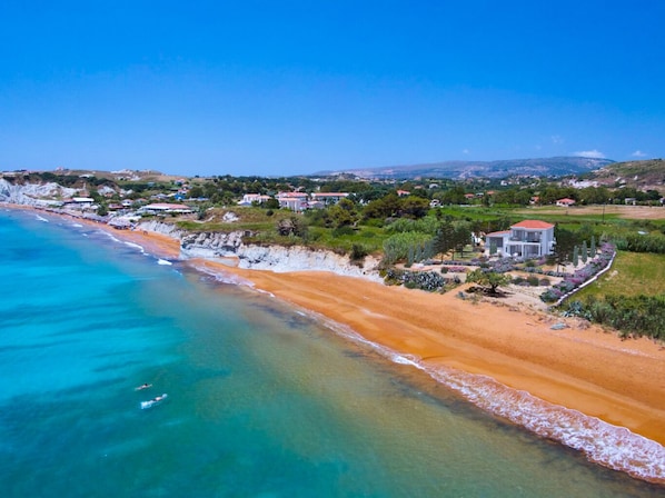 A coastal area with a sandy beach, a clear blue sea, and a few buildings with red roofs. - Grand Blue Beach Apartments & Suites (Kefalonia)