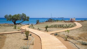 Ein hölzerner Steg schlängelt sich durch eine Wüstenlandschaft mit spärlicher Vegetation, der zu einem Strand mit klarem, blauem Wasser und fernen Inseln führt.
