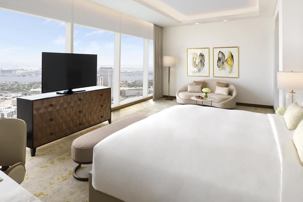 A modern hotel room with a large bed, a TV on a wooden dresser, a sofa, and a view of the city.