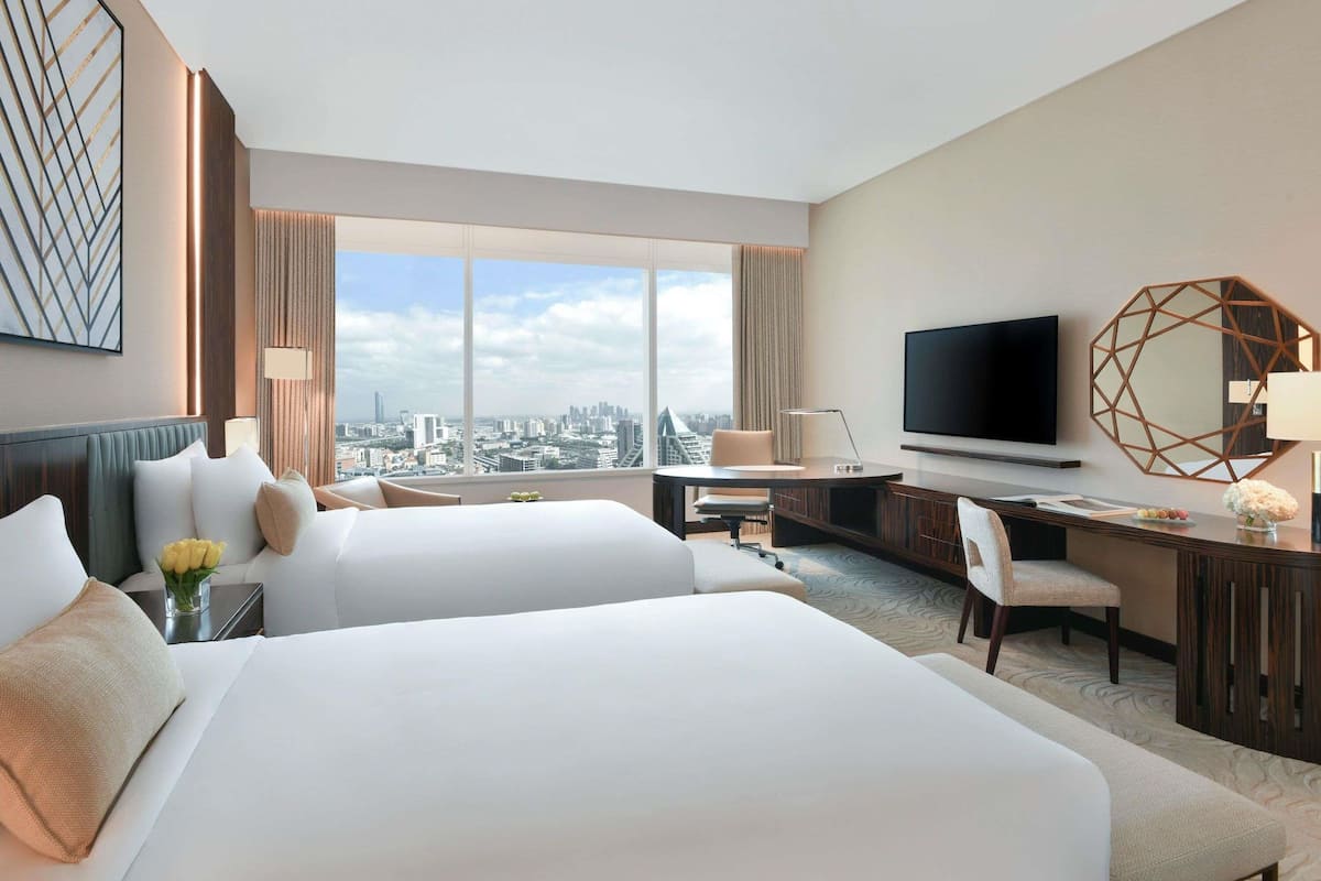 A hotel room with two beds, a desk, a TV, and a large window with a city view.