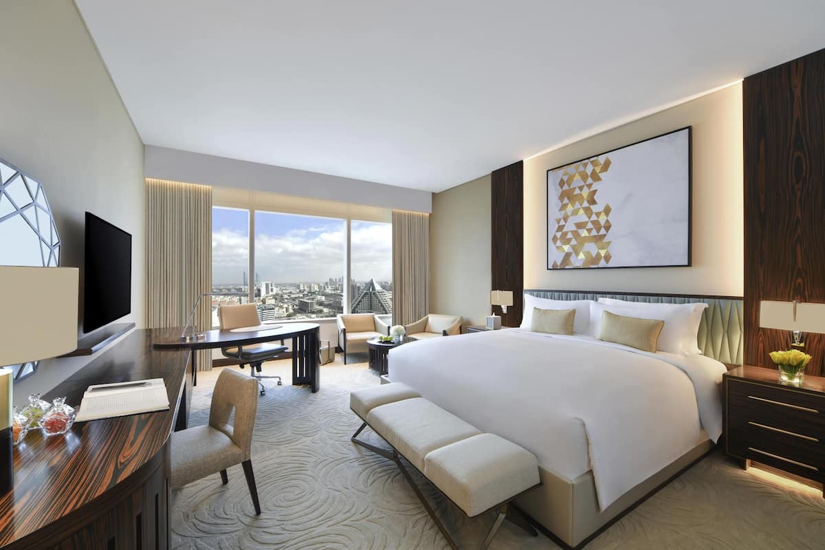 Habitación de hotel con una cama grande, un escritorio con silla, televisor y vistas a la ciudad a través de amplios ventanales.