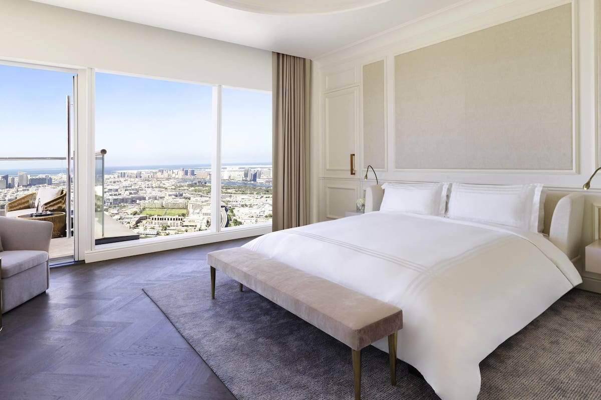 A spacious bedroom with a large bed, a bench, a nightstand, and a view of a cityscape through a large window.