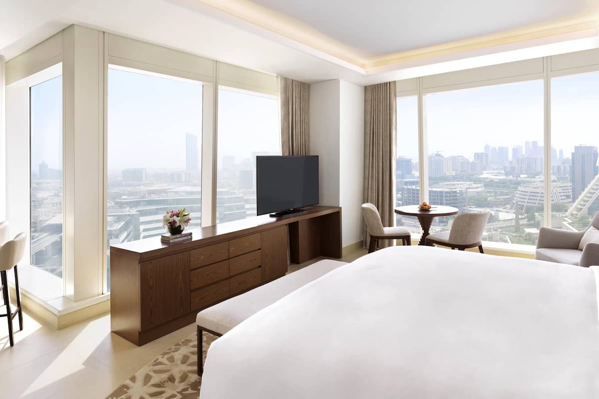 A modern hotel room with a large bed, a TV on a wooden console, a desk with a chair, and a view of the cityscape through large windows.
