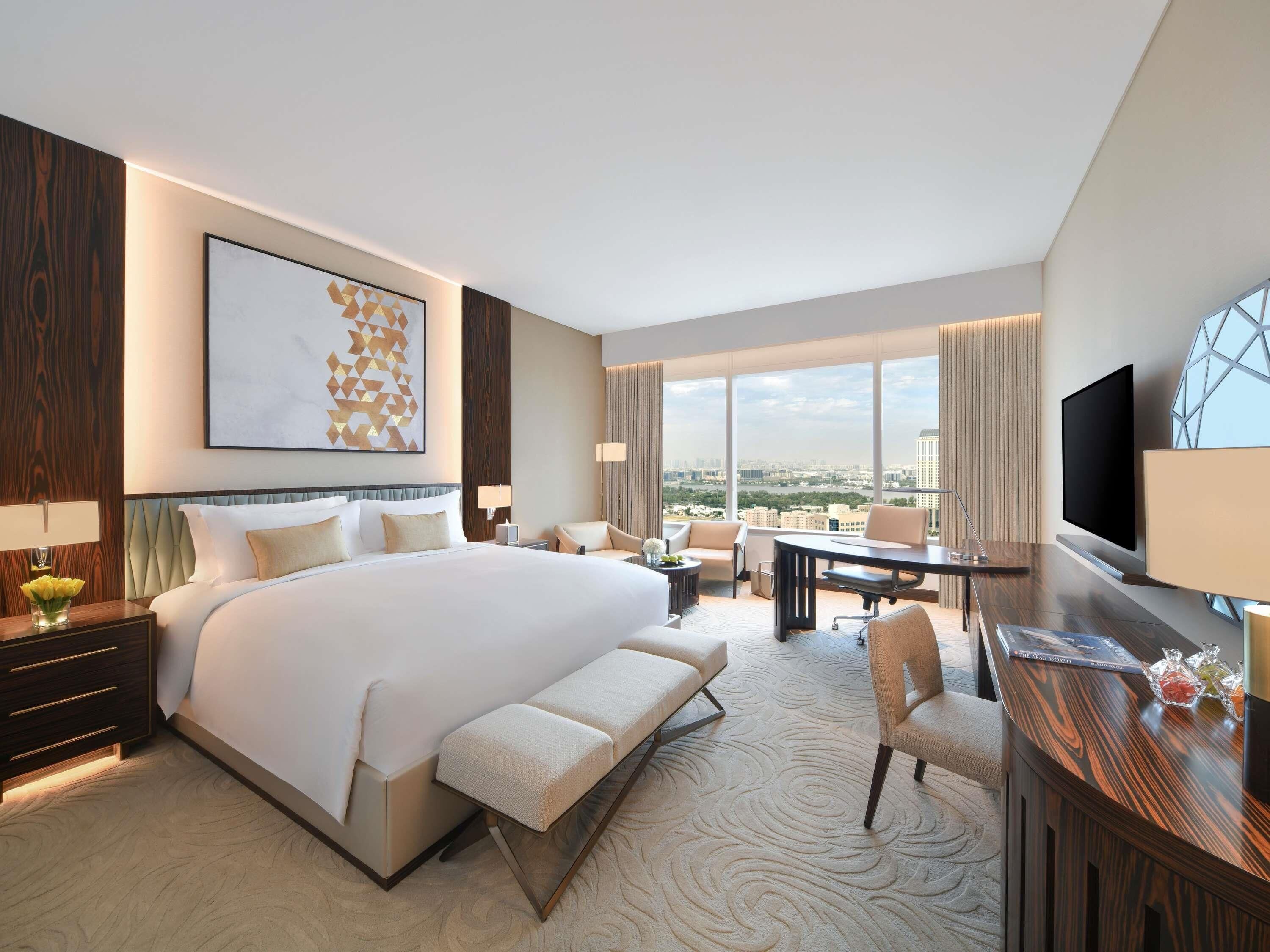 A modern hotel room with a large bed, a desk, a chair, a TV, and a city view.