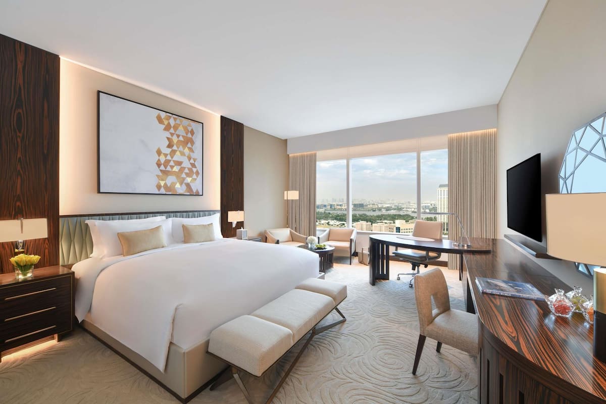 A modern hotel room with a large bed, a desk, a chair, a TV, and a city view.