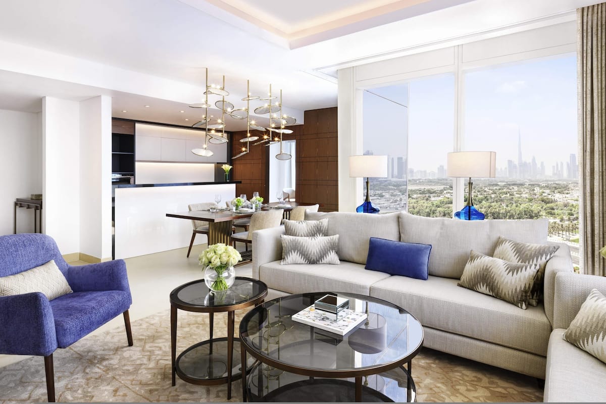 A modern living room with a sofa, armchairs, and a coffee table. There is a dining area with a table and chairs, and a kitchen in the background.