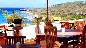 A patio with wooden tables and chairs, a stone basin, and a view of the ocean and hills. - Oceanview B&B Curacao (Soto)
