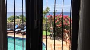 A view through a window with a pool and lounge chairs, overlooking a beach and pink flowers. - Oceanview B&B Curacao (Soto)