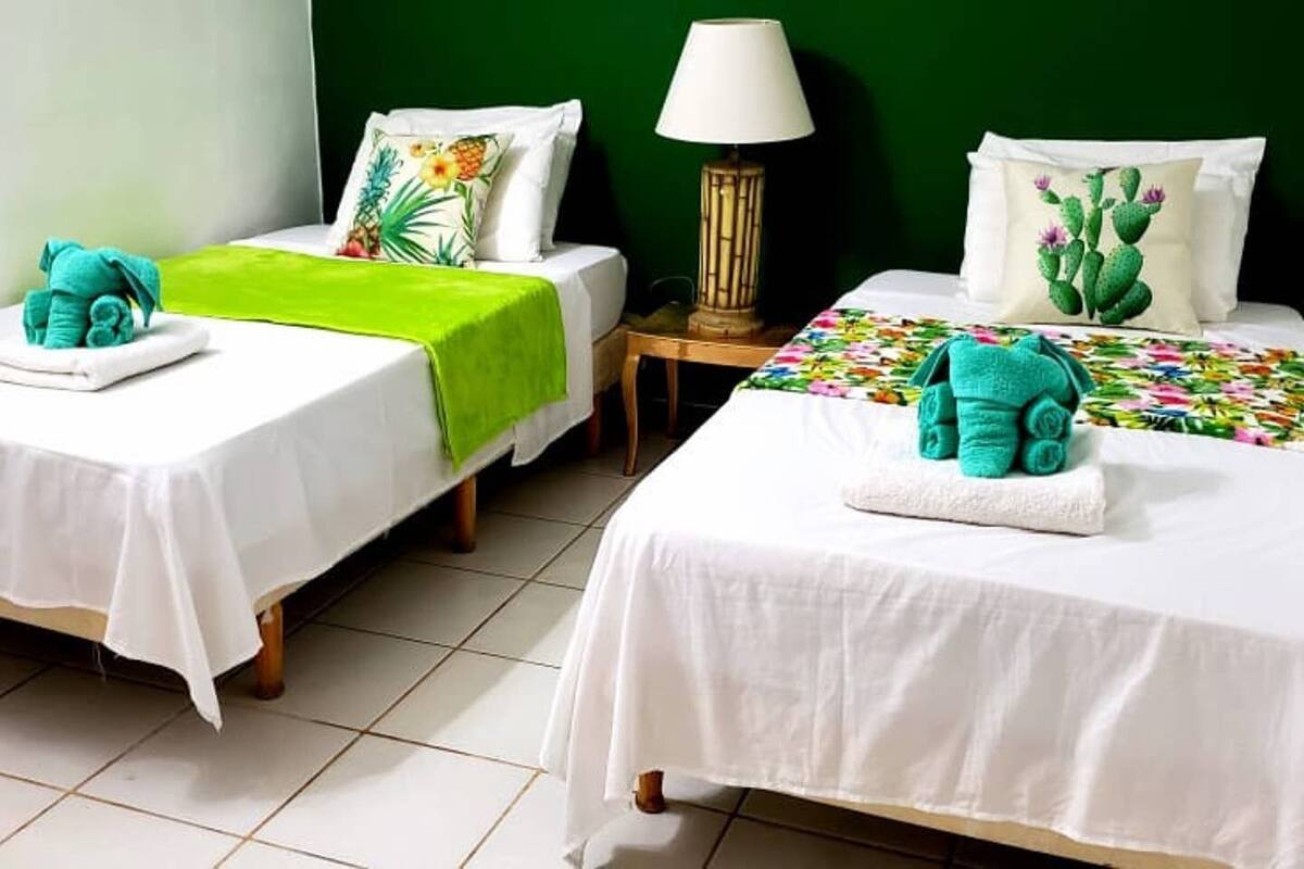 A hotel room with two beds, each with a white bedspread and a green towel. There is a lamp on a wooden nightstand between the beds.