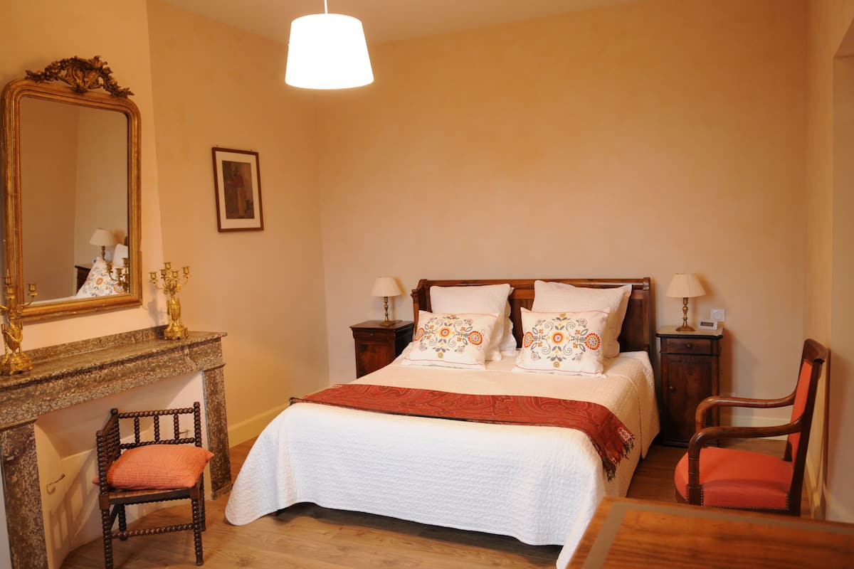 A bedroom with a bed, two bedside tables, a nightstand, a chair, a dresser, a mirror, and a hanging light fixture.