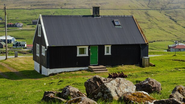 A black house with a green door, surrounded by rocks and grass, set against a cloudy sky and hills in the background. - The Real Faroese Experience (Skalavik)