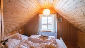 A cozy attic bedroom with a wooden sloped ceiling, a single bed with white bedding, a window with curtains, and a hanging light fixture. - The Real Faroese Experience (Skalavik)